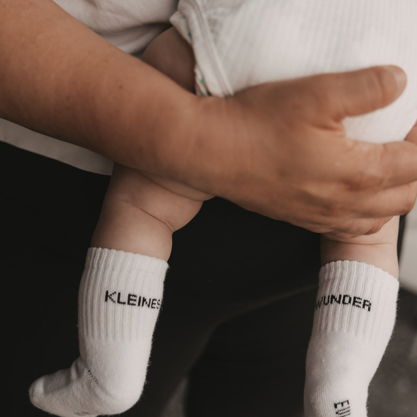 Baby wird gehalten, trägt weiße Socken mit „Kleines Wunder“ Schriftzug, liebevolle Aufnahme