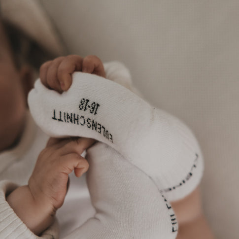 Baby greift nach Fuß, trägt weiße Socken mit Schrift „Kleines Wunder“, Nahaufnahme
