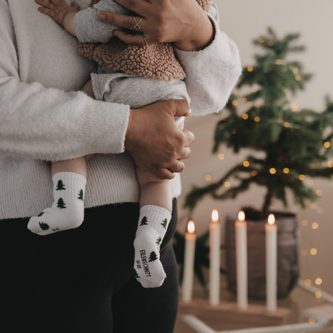 Baby mit Tannen Socken auf dem Arm, weihnachtliche Szene mit Kerzen und Tannenbaum im Hintergrund