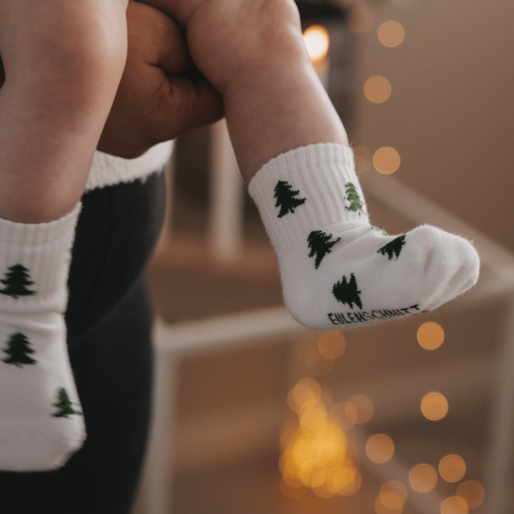 Nahaufnahme Babyfuß mit weißen Socken und Tannenmotiv, warme Lichter im Hintergrund