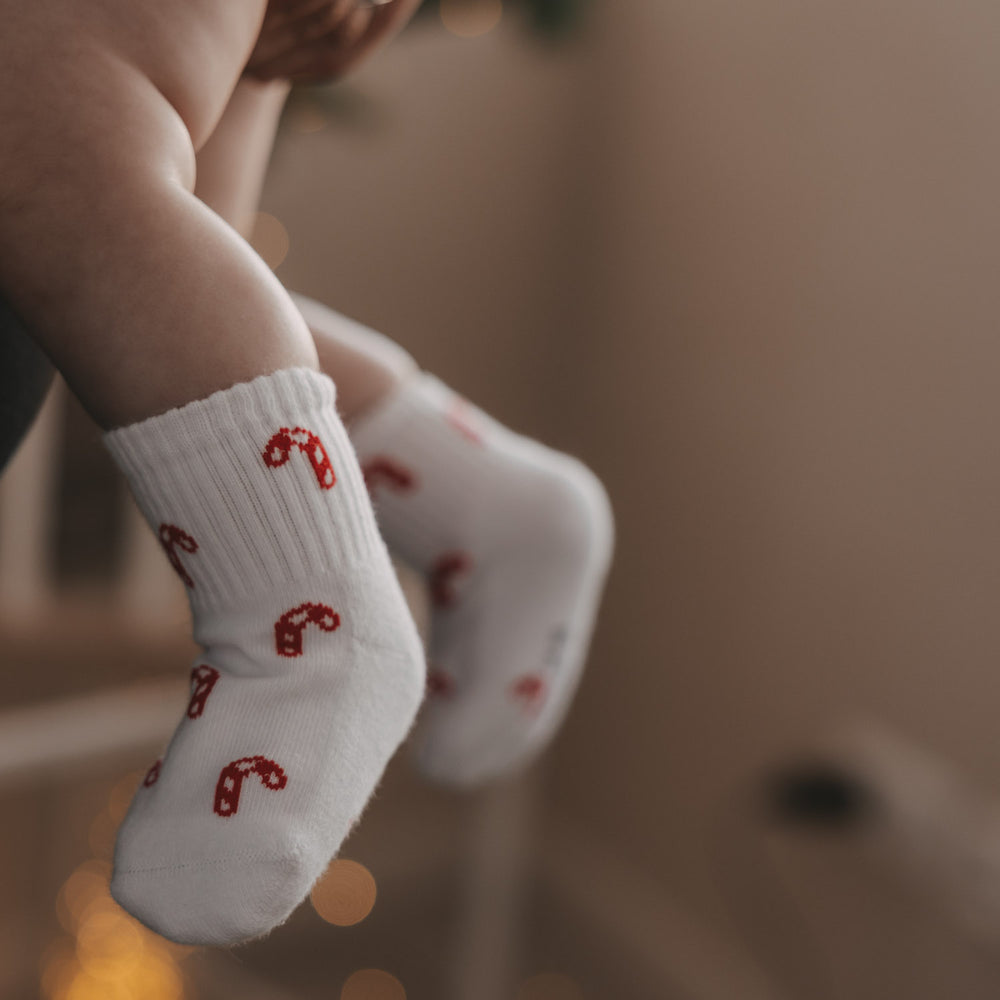 Nahaufnahme Babyfüße mit weißen Socken und roten Zuckerstangen, warmes Licht im Hintergrund