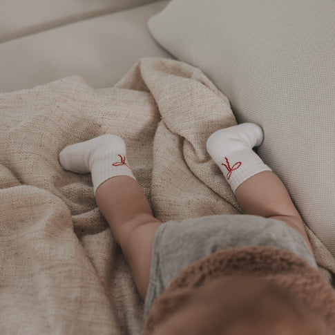 Baby liegt mit weißen Socken und roter Schleife Motiv auf Decke, ruhige und natürliche Szene