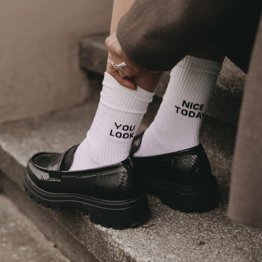 Person sitzt auf einer Treppe und trägt weiße Socken mit der Aufschrift „YOU LOOK“ und „NICE TODAY“ – kombiniert mit eleganten schwarzen Loafern für einen stilvollen und charmanten Look.