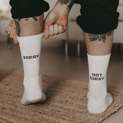 Person mit Tätowierungen trägt weiße Socken mit den Schriftzügen „SORRY“ und „NOT SORRY“ – humorvoller Kontrast auf gemütlichem Teppich.