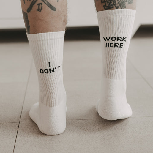Weiße Socken mit schwarzem Schriftzug „I DON’T“ und „WORK HERE“ an tätowierten Beinen – von hinten auf hellen Boden fotografiert.