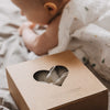 Verpackung des Musselintücher-Sets mit Herzfenster im Vordergrund, während ein Baby im Hintergrund auf einer weichen Decke liegt. Die Szene vermittelt Wärme und Geborgenheit im Babyalltag.