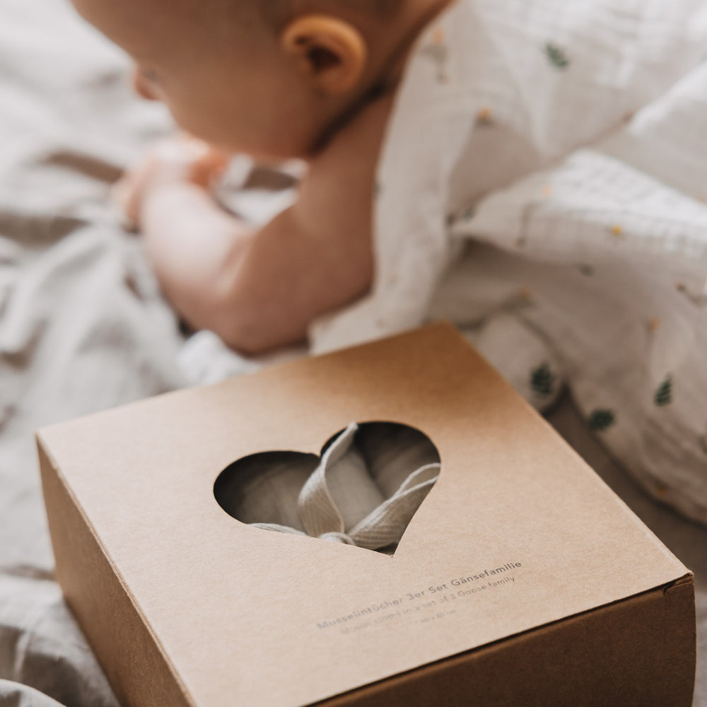 Verpackung des Musselintücher-Sets mit Herzfenster im Vordergrund, während ein Baby im Hintergrund auf einer weichen Decke liegt. Die Szene vermittelt Wärme und Geborgenheit im Babyalltag.