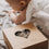 Verpackung des Musselintücher-Sets mit Herzfenster im Vordergrund, während ein Baby im Hintergrund auf einer weichen Decke liegt. Die Szene vermittelt Wärme und Geborgenheit im Babyalltag.