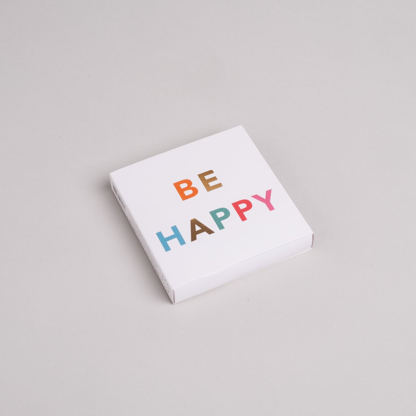 Streichholzschachtel mit bunter Aufschrift BE HAPPY auf weißem Karton, moderne Geschenkidee.