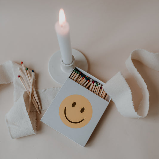 Geöffnete Smiley-Streichholzschachtel neben brennender Kerze und losen Streichhölzern.