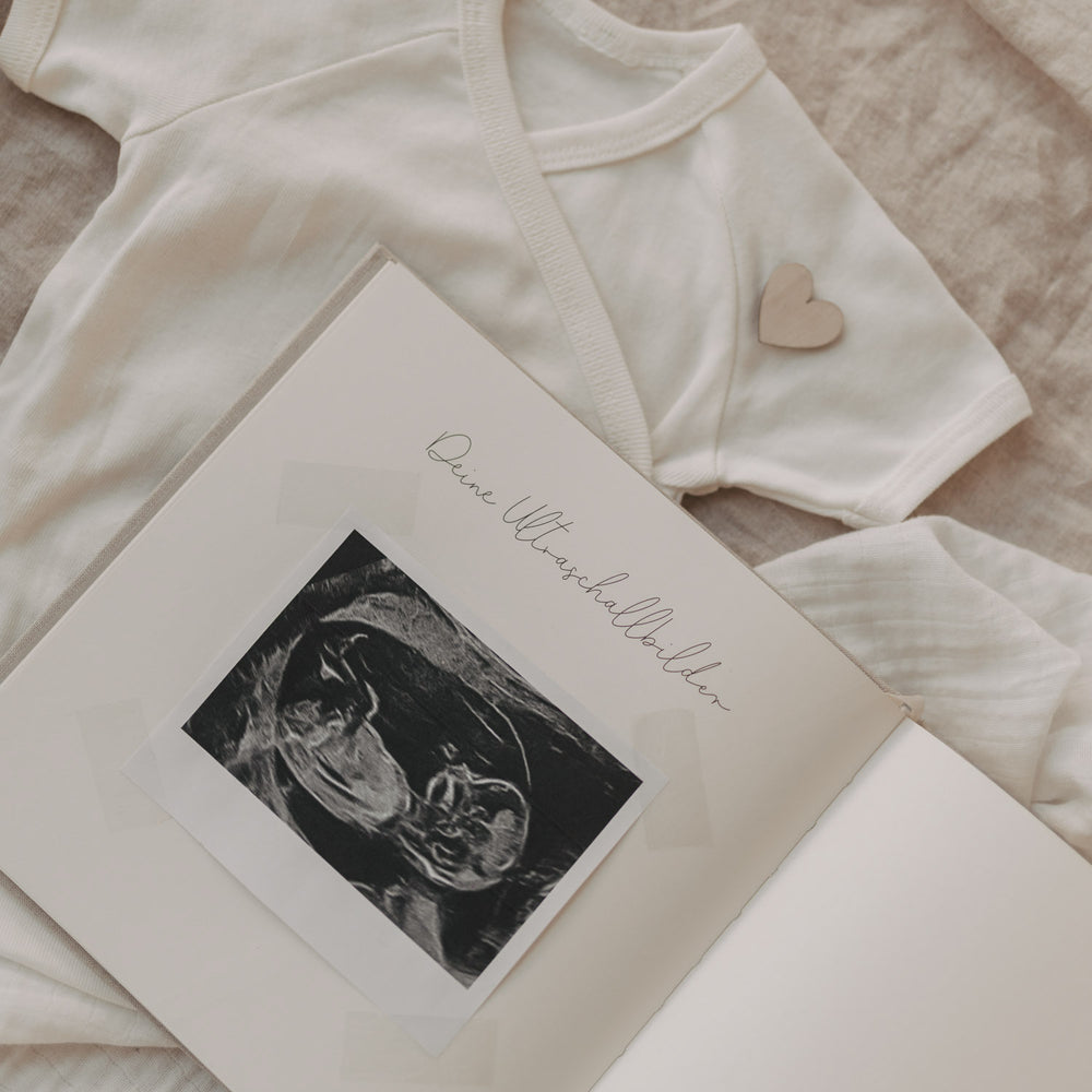Geöffnetes Schwangerschaftstagebuch mit eingeklebtem Ultraschallbild auf Babybody liegend.