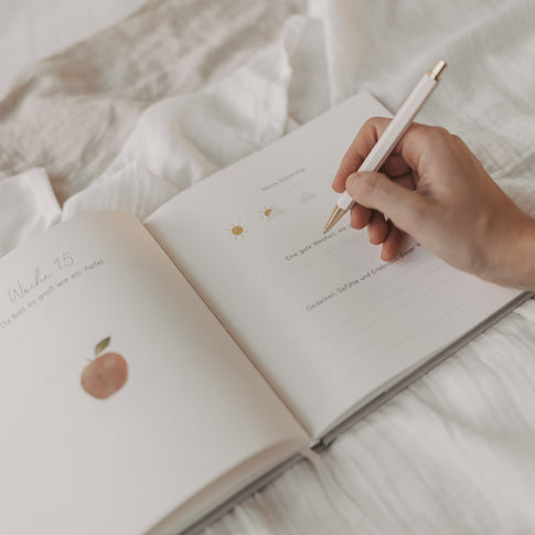 Hand schreibt ins Schwangerschaftstagebuch auf Seite zur 15. Woche mit Illustrationen.