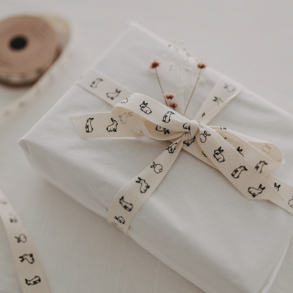 Weiße Geschenkbox mit Kaninchen-Geschenkband und Trockenblumen auf hellem Tisch.