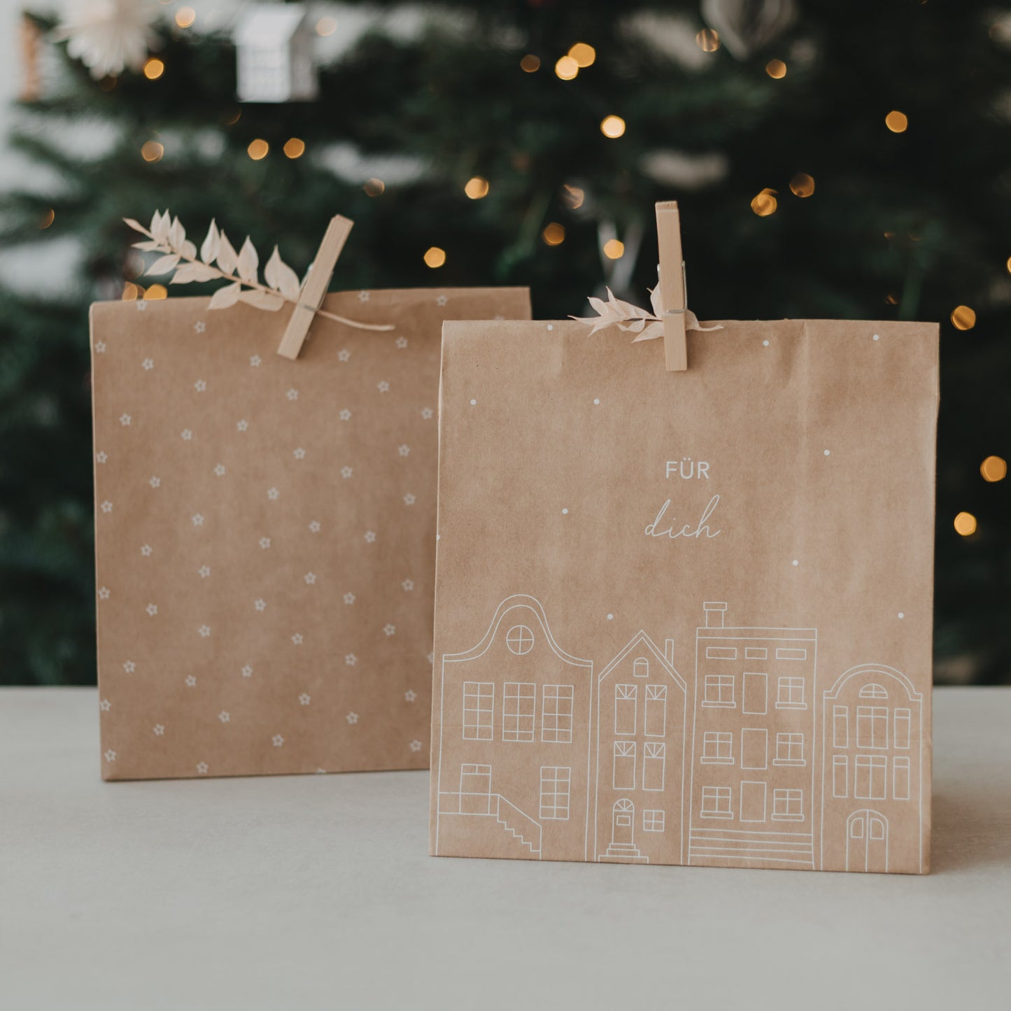 Geschenktüte aus Kraftpapier mit weißer Häuserillustration und Schriftzug „Für dich“ vor Weihnachtsbaum