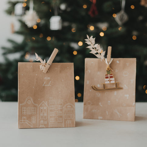Geschenktüte aus Kraftpapier mit „Für dich“ und Häuser-Motiv neben weihnachtlich dekorierter Tüte